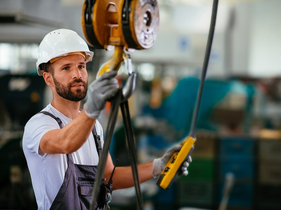 Das Bild zeigt einen Mitarbeiter von seitlich rechts im Porträt, der ein weißes T-Shirt, eine dunkle Latzhose, einen weißen Helm und graue Arbeitshandschuhe trägt, in einer Werkshalle. In seiner linken Hand hält er eine Kranflasche. In der rechten Hand hält einen vom Hallenkran außerhalb des Bildes herabgelassenen Kranhaken, an dem ein schwarzes Band angeschlagen ist. Der Mitarbeiter schaut auf den Kranhaken und scheint ihn zu prüfen. Link zur vergrößerten Darstellung des Bildes.