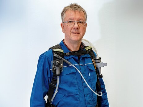 Olaf Binder frontal im Porträt mit direktem Blick zum Betrachter. Er trägt einen Blaumann und darüber einen Brustgurt mit verschiedenem Mess-Equipment. Links ein zylinderförmiger Messkopf mit Schlauch, der über die Schulter auf den Rücken führt. Rechts ein langes, dünnes zylinderförmiges Gerät, das wie ein Mikrofon aussieht. Davon führt über die Brust auf ein weißer Schlauch auf die linke Seite von Herrn Binder. Unter dem rechten Gerät hängt ein weiteres, das aussieht wie eine Spritzpistole. Unterhalb davon führt ein dritter Schlauch an der Hüfte entlang auf Herrn Binders Rücken. Link zur vergrößerten Darstellung des Bildes.