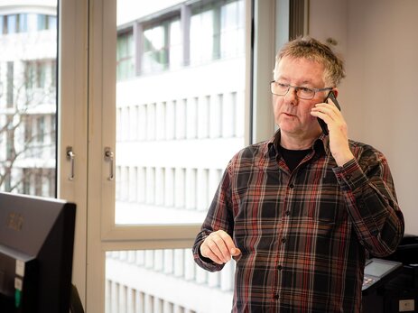 Olaf Binder frontal im Porträt, der sich ein Telefon an das linke Ohr hält und auf einen Bildschirm am unteren linken Bildrand schaut. Hinter ihm ist ein Fenster in dem ein Bürogebäude zu sehen ist. Link zur vergrößerten Darstellung des Bildes.