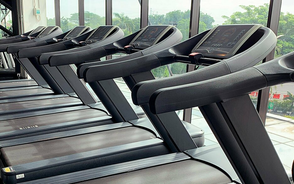 Das zeigt eine Reihe Laufbänder in einem Fitnessstudio vor einem großen Panoramafenster, hinter dem Natur und Bäume zu sehen sind. Link zum Artikel.