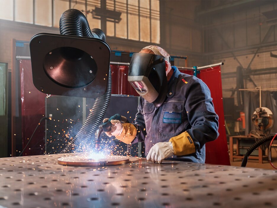 Das Bild zeigt einen Mitarbeiter in einer Werkstatt an einem Schweißertisch. Er trägt ein Schweißerschutzvisier und Schutzhandschuhe. In der rechten Hand hält er ein Schweißgerät, mit dem er gerade ein Stück Metall schweißt, das auf dem Tisch liegt. Über seiner rechten Schulter ist der Trichter eines großen, schwarzen Absaugschlauchs zu sehen, der nach unten führt.. Link zur vergrößerten Darstellung des Bildes.