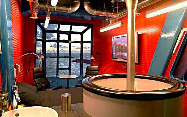 Das Bild zeigt den Blick in das Zimmer eines zum Hotel umgebauten Hafenkrans. Die Wände sind in rot-blau gehalten. Im Vordergrund hängt ein Waschbecken, dahinter sind ein Bett, zwei Stühle und ein kleiner Tisch zu sehen. Gegenüberliegend ist ein Fenster mit dem Ausblick auf die Nordsee. Link zum Artikel.