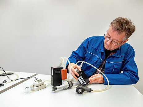 Olaf Binder sitzt an einem Tisch und schaut auf das Display eines schwarzen Kastens, der vor ihm steht. Der Kasten ist per Schlauch mit einem Messkopf verbunden, der wie eine Garten-Spritzpistole verbunden ist. Daneben ist ein zweiter schwarzer Kasten, der per Schlauch mit einem Messkopf verbunden ist, der wie eine metallische Flasche aussieht. Daneben steht ein weißes Gerät mit Display, in dem die Zahl „1030“ steht. Link zur vergrößerten Darstellung des Bildes.