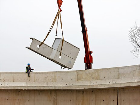 Das Bild zeigt ein Betonteil, das schief von einem Kran transportiert wird. Im Vordergrund sitzt ein Mitarbeiter ungesichert in der Höhe auf einem anderen Betonteil und gibt dem Kranführer Handzeichen. Es sieht so aus, als ob das schief hängende Teil über den Kopf des Mitarbeiters transportiert wird. Link zur vergrößerten Darstellung des Bildes.