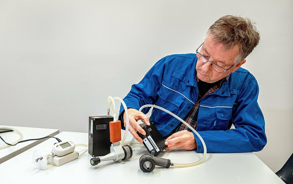 Olaf Binder sitzt an einem Tisch und schaut auf das Display eines schwarzen Kastens, der vor ihm steht. Der Kasten ist per Schlauch mit einem Messkopf verbunden, der wie eine Garten-Spritzpistole verbunden ist. Daneben ist ein zweiter schwarzer Kasten, der per Schlauch mit einem Messkopf verbunden ist, der wie eine metallische Flasche aussieht. Daneben steht ein weißes Gerät mit Display, in dem die Zahl „1030“ steht. Link zum Artikel.