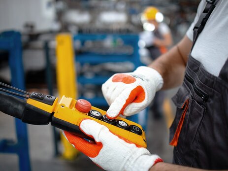 Das Bild zeigt die Hände eines Mitarbeiters in einer Arbeitshalle. Er trägt Handschuhe und hält eine Kranflasche in der linken Hand. Mit dem rechten Zeigefinger drückt der Mitarbeiter auf einen Knopf auf der Kranflasche. Link zur vergrößerten Darstellung des Bildes.