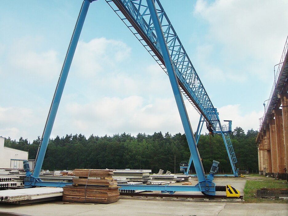 Das Bild zeigt einen großen blauen Portalkran auf einem Betriebsgelände von der Seite. Links ist eine Halle zu sehen, im Hintergrund Bäume und im Vordergrund liegen Holzplatten neben den Laufschienen des Krans. Link zur vergrößerten Darstellung des Bildes.