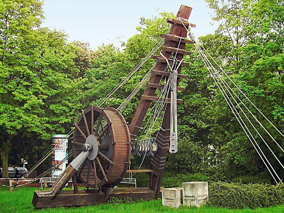 Das Bild zeigt einen alten Römerkran mit Tretmühle im Grünen. Dabei ist die Hebevorrichtung schräg nach oben gerichtet und mit Seilen gesichert. Neben der Hebevorrichtung ist ein großes, hölzernes Rad mit Holzspeichen angebracht. In diesem sind früher sogenannte Windenknechte gelaufen, um den Kran zu bewegen und Lasten anzuheben. Link zur vergrößerten Darstellung des Bildes.