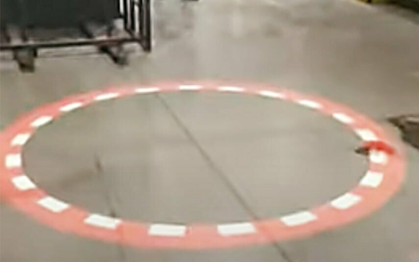 Das Bild zeigt den Blick auf den Boden einer Halle. Zu sehen ist ein leuchtender rot-weißer Kreis auf dem Boden. Es sieht aus, wie ein warnendes Flatterband. Der Kreis wird von oben von einem Kran auf den Boden projiziert. Er warnt Fußgänger vor dem Gefahrenbereich. Link zum Artikel.