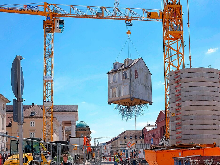 Das Bild zeigt den Blick auf eine Fußgängerzone in einer Stadt. An den Seiten des Bildes sind 2 gelbe Kräne zu sehen. An einem der Kräne hängt ein Haus, das unten viele Wurzeln hat. Drumherum auf dem Boden sind viele Bauzäune aufgestellt. Es ist eine Kunstinstallation, die auf Vertreibung und Entwurzelung aufmerksam machen soll. Link zur vergrößerten Darstellung des Bildes.