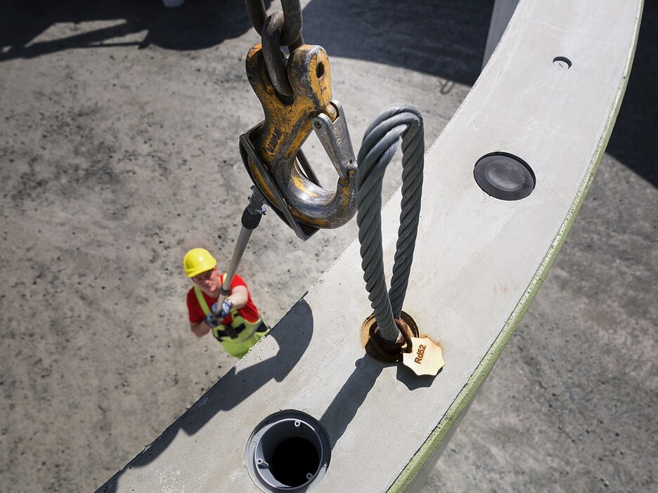 Zu sehen ist ein Mitarbeiter von oben, der neben einem großen, anzuschlagenden Betonteil steht. Er hält einen Stangenaufsatz mit Kelle in der Hand, mit dem er den Kranhaken oben führt und in die Öse des Betonfertigteils drückt. Für das Entfernen der Haken wird später dann ein anderer Aufsatz an der Stange verwendet. Link zur vergrößerten Darstellung des Bildes.